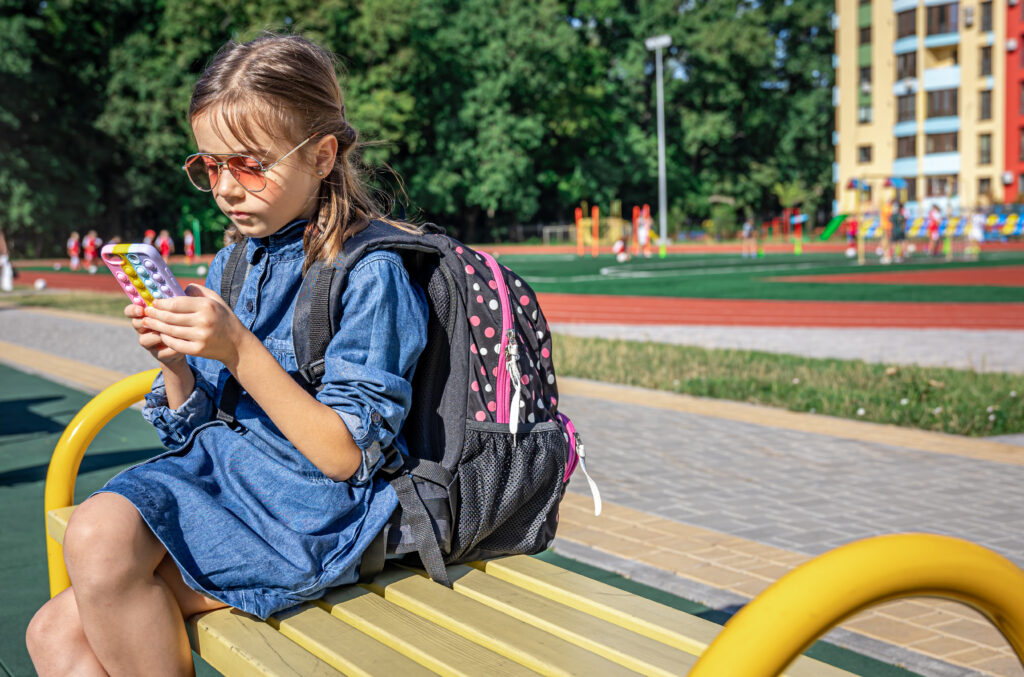 A Primary School Student With A Backpack Uses A Smartphone Sitting Near The School